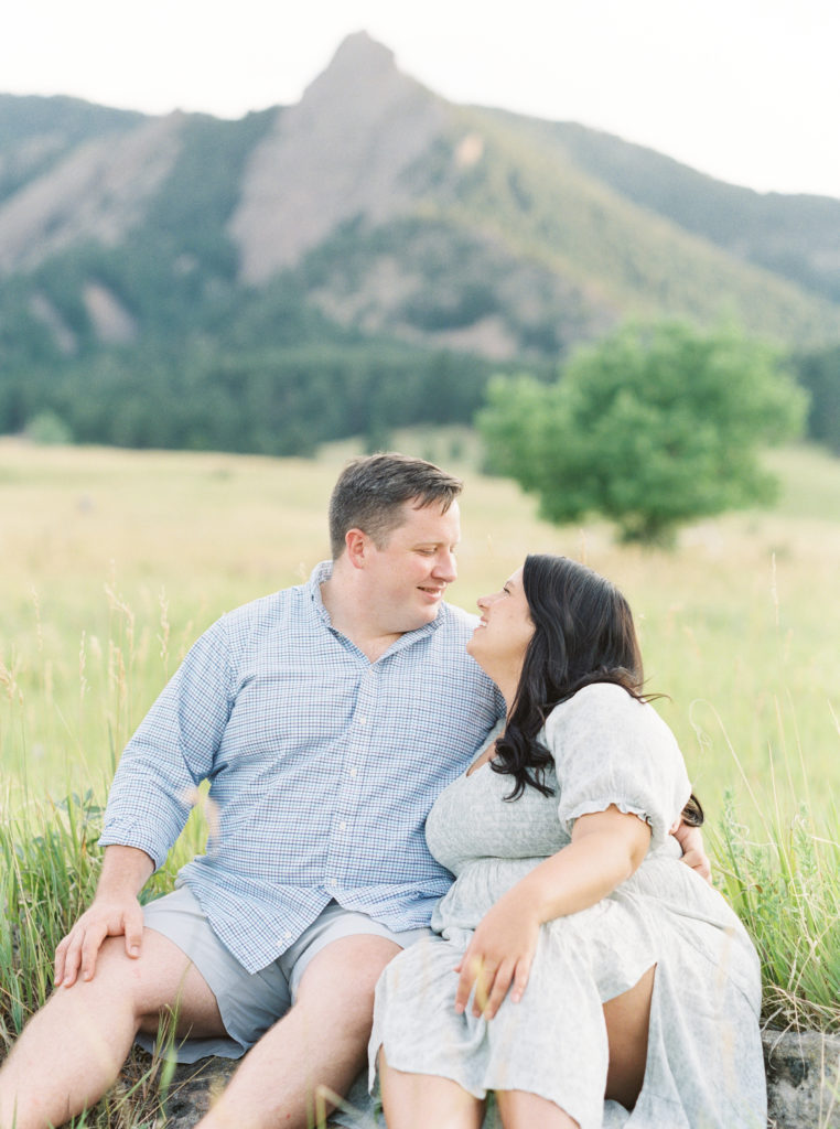 Couple smiles on camera with Flatirons in the background for mountainside engagement photos in Colorado